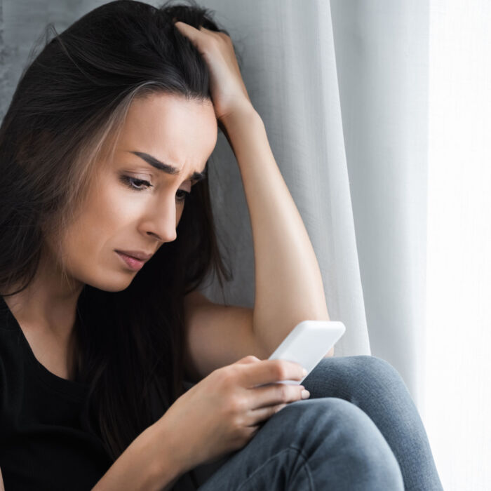 disappointed girl using smartphone while sitting by window at home and holding hand on head